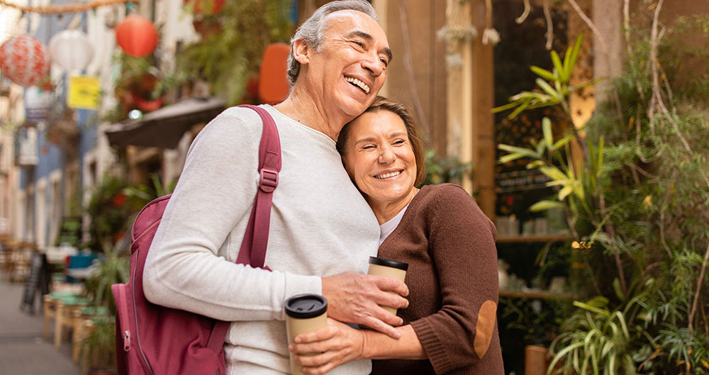 Happy Vacation Travel. Cheerful Mature Spouses Enjoying Their Lisbon Getaway, Hugging Posing With Backpack And Paper Coffee Cups Outside. Tour Offer For Retired People Concept